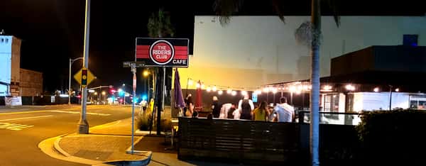 The Riders Club Café at night with string lights illuminating a lively outdoor patio filled with diners on a city street corner in San Clemente, California.
