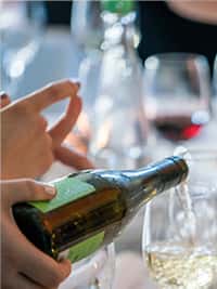 Close-up of a wine bottle being poured into a glass during a lively dining event, with blurred wine glasses and ambient lighting in the background, capturing an upscale and social restaurant atmosphere.