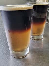 Two pints of layered black and tan beer, showcasing a striking contrast between dark stout and golden ale, served cold on a stainless steel counter at The Riders Club Café.