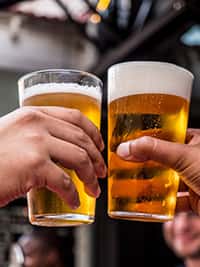 Close-up of two people clinking glasses of golden craft beer in a celebratory toast at The Riders Club Café, capturing the lively and social ambiance of the venue.
