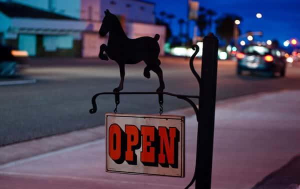 Evening street view featuring an illuminated "OPEN" sign hanging beneath a silhouette of a rearing horse, signaling that The Riders Club Café is open for business.