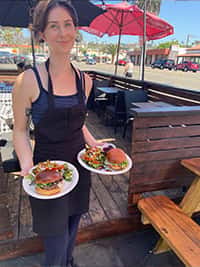 Outdoor patio dining scene featuring a server in a black apron holding two vibrant plates with gourmet burgers and colorful side salads at The Riders Club Café, under red umbrellas with a lively street view in the background.
