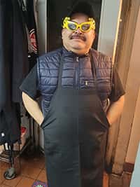 Restaurant staff member at The Riders Club Cafe wearing a black apron, navy padded vest, and festive yellow novelty glasses, smiling playfully in a kitchen doorway.