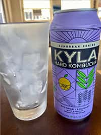 Frosted glass filled with ice next to a vibrant purple can of KYLA Hard Kombucha, Lavender Lemonade flavor from the Sunbreak Series, on a wooden table in a sunlit setting.