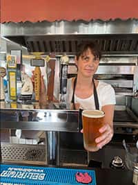 Bartender serving a freshly poured pint of craft beer from a tap at The Riders Club Café, with multiple beer tap handles in the background inside a casual, open-kitchen bar setting.