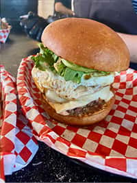Gourmet breakfast burger with a fried egg, melted cheese, fresh lettuce, and a juicy beef patty on a toasted brioche bun, served on red-and-white checkered paper at The Riders Club Café.
