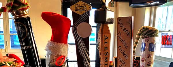 Close-up of a festive beer tap lineup at a craft beer bar, featuring handles from Karl Strauss, North Coast Brewing, Alesmith, and Docent Brewing, some adorned with holiday hats and decorations.