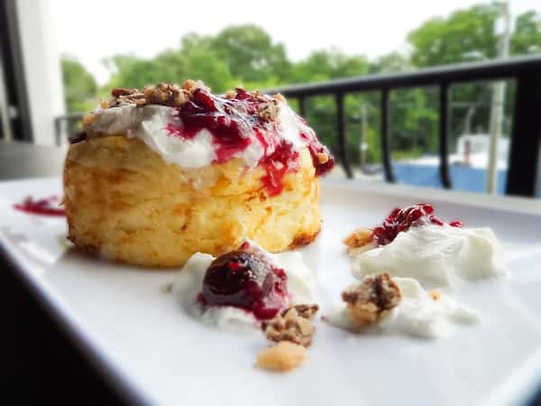 Mini cheesecake dessert topped with whipped cream, raspberry compote, and crunchy crumble, plated on a white dish with an outdoor patio view.