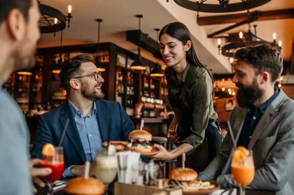 Friendly server delivering gourmet burgers to a group of diners in a warm, modern restaurant, with craft cocktails and a lively casual dining atmosphere