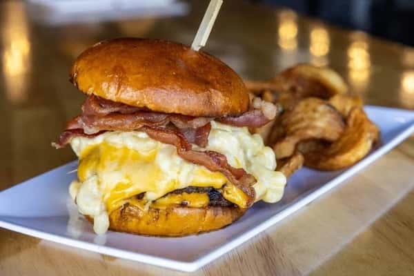 Stacked breakfast burger with melted cheddar, creamy scrambled eggs, and crispy bacon on a glossy brioche bun, served with kettle-style potato chips in a cozy restaurant setting