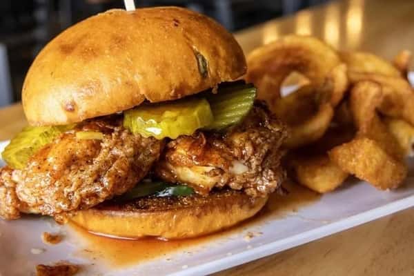 Crispy fried chicken sandwich on a toasted brioche bun with pickle slices and spicy sauce, served with golden onion rings on a wooden table in a casual restaurant setting.