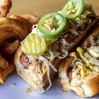 Grilled sausage sandwich topped with sauerkraut, pickle slices, and jalapeños on a toasted bun, served with crispy potato sides in a casual diner setting.