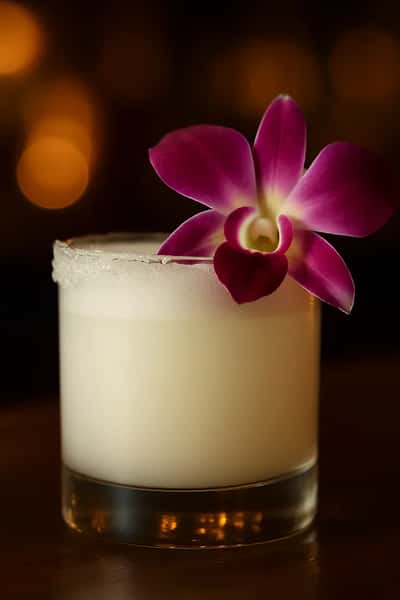 Creamy tropical cocktail in a short glass garnished with a purple orchid, set against a warm, dimly lit bar ambiance.