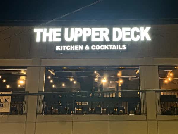 Exterior view of The Upper Deck Kitchen & Cocktails restaurant at night, featuring illuminated signage and a rooftop patio with string lights for an inviting nightlife dining experience