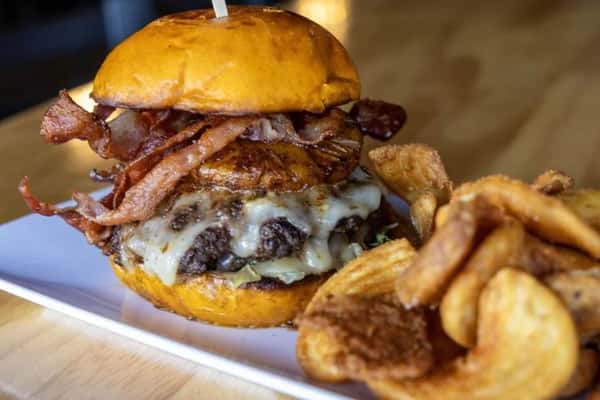Gourmet bacon cheeseburger with melted cheddar, crispy bacon strips, and a toasted brioche bun, served with golden house-made potato chips in a casual restaurant setting