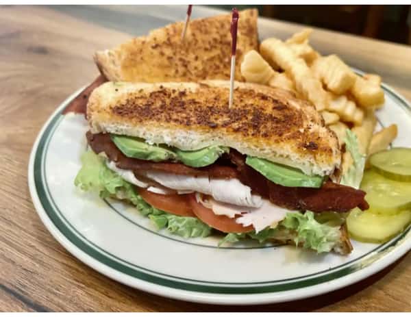 Grilled turkey club sandwich with avocado, bacon, lettuce, and tomato, served with crinkle-cut fries and pickles on a white plate.