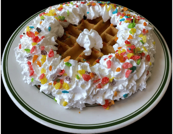 Golden waffle topped with whipped cream and colorful Fruity Pebbles cereal, served on a white plate with green trim.