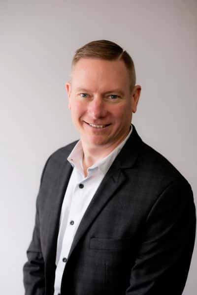 A man in a dark suit jacket and white shirt smiles warmly, posing against a neutral background for a professional portrait.