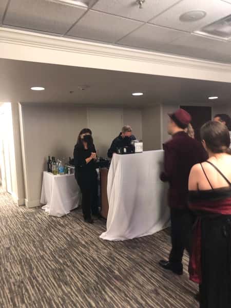 Guests in formal attire wait at a bar station with masked bartenders serving drinks at an indoor catered event.