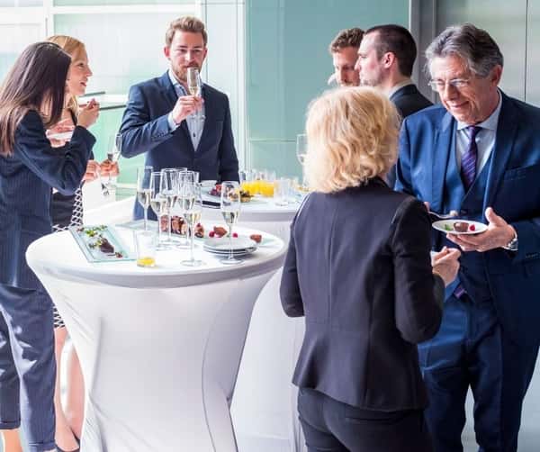 Business professionals networking at a cocktail reception with champagne glasses, hors d'oeuvres, and high-top tables.