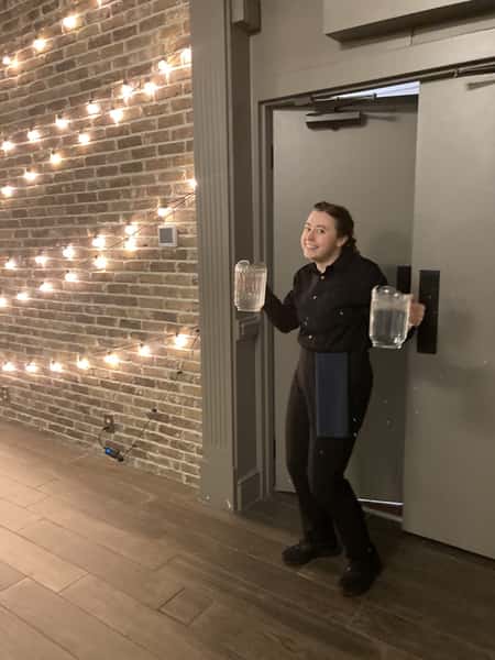 Smiling server in black uniform carrying two pitchers of water, standing by string lights on a brick wall in an event venue.