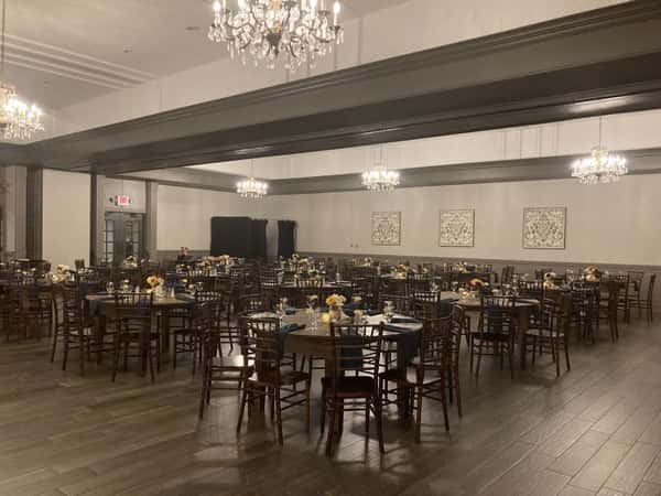 Elegant banquet hall with chandeliers, round tables, dark wooden chairs, and floral centerpieces arranged for a formal event.