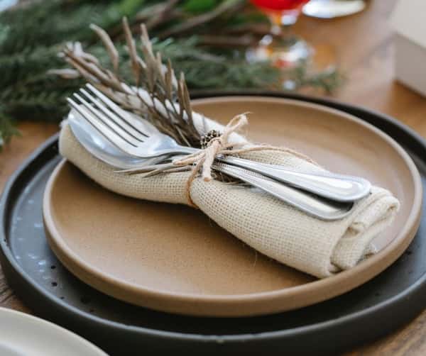 Rustic table setting with neutral-toned plates, cutlery wrapped in burlap, tied with twine, and decorated with dried foliage.
