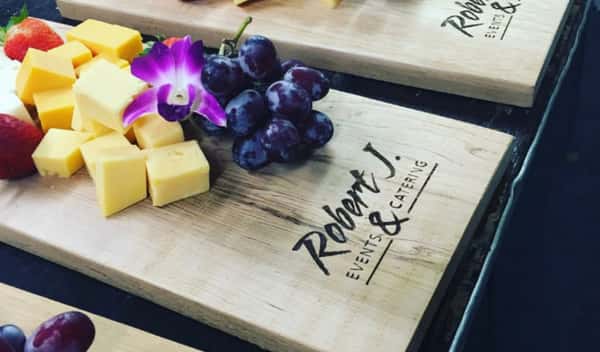 A cheese and fruit platter on a wooden board branded with "Robert J. Events & Catering," featuring cubes of cheddar cheese, grapes, strawberries, and a purple orchid for garnish.
