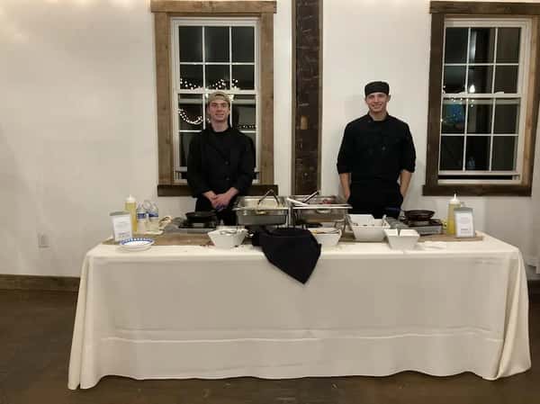 Two chefs in black uniforms stand behind a catering station with chafing dishes and serving bowls in a rustic venue.