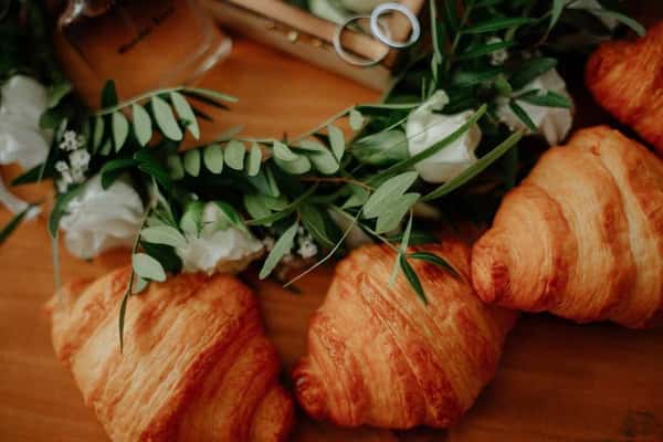 This image shows a few golden, flaky croissants placed next to a decorative arrangement of white flowers and green leaves. Above the croissants, a pair of wedding rings is visible, suggesting the image might be styled for a wedding or celebratory breakfast/brunch scene.