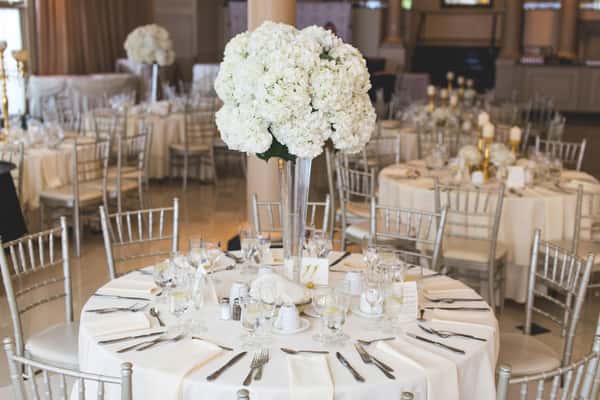 Elegant round banquet table with white linens, silver chairs, and a tall centerpiece of white hydrangeas in a clear vase.