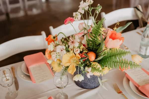Elegant table setting with pink napkins, menus, glassware, and a vibrant floral centerpiece with greenery and colorful blooms.