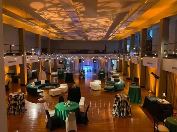 Elegant event hall with dramatic ceiling lighting, round tables with green covers, modern lounge seating, and string lights above.