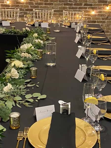 Elegant banquet table with black linens, gold chargers, black napkins, greenery centerpiece, candles, and name cards under string lights.