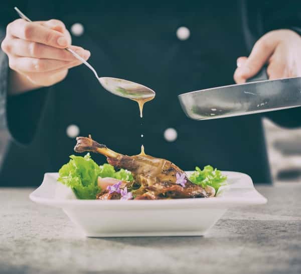 Chef pouring sauce over a plated roasted duck leg with fresh greens and edible flowers for a fine dining presentation.