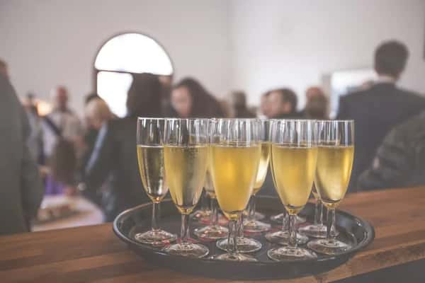 Tray of champagne flutes filled with sparkling wine on a wooden counter at a social gathering with blurred guests in the background.