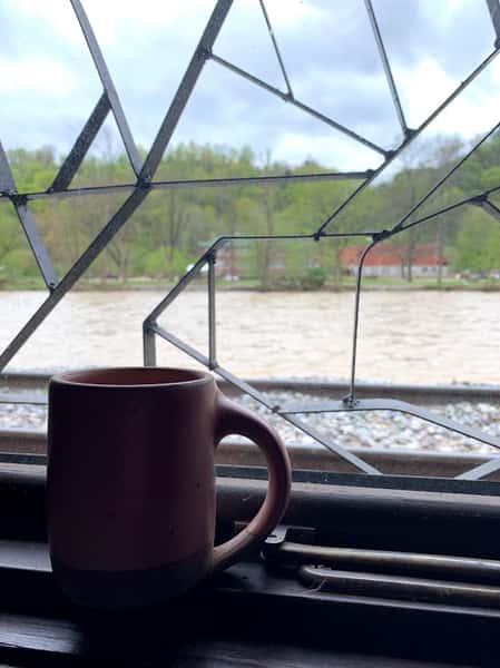A brown mug sits on a window sill. Outside the window is a fast-flowing river and the opposite shore on a cloudy day.