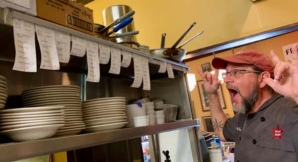 A busy chef in a busy kitchen. Restaurant tickets hang above a shelf of plates, with "Beef Tender" and "Emily" written on them.