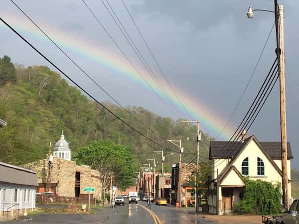 A rainbow ends in a small town, appearing to touch the top of a white building. The wet street reflects buildings, power lines, and green hills in the background.