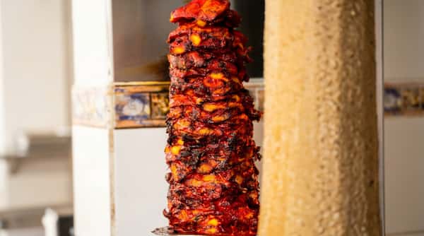 Vertical rotisserie of al pastor meat, a large cone of reddish-brown marinated pork with visible charred pieces.