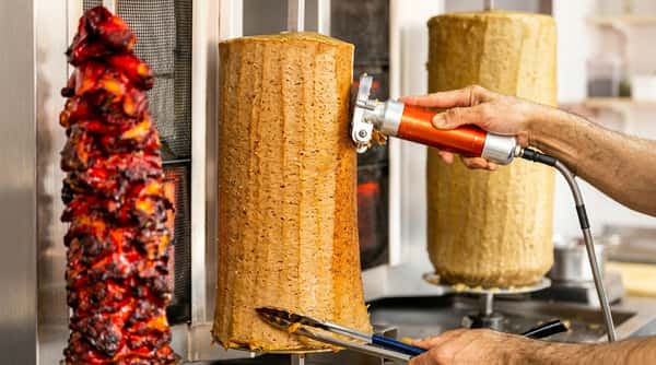 A hand using an electric knife to shave meat from a large vertical doner kebab spit.