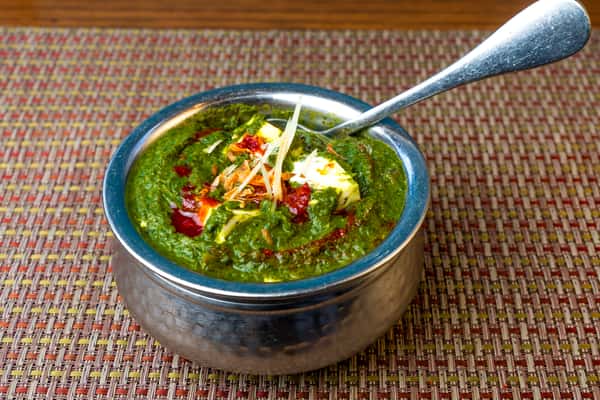 Palak paneer — creamy spinach curry with paneer cubes, topped with chili oil, butter, and julienned ginger in a traditional metal bowl.
