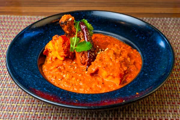 Chicken curry in a rich spiced tomato gravy with dried chili and cilantro garnish, served on a blue plate.