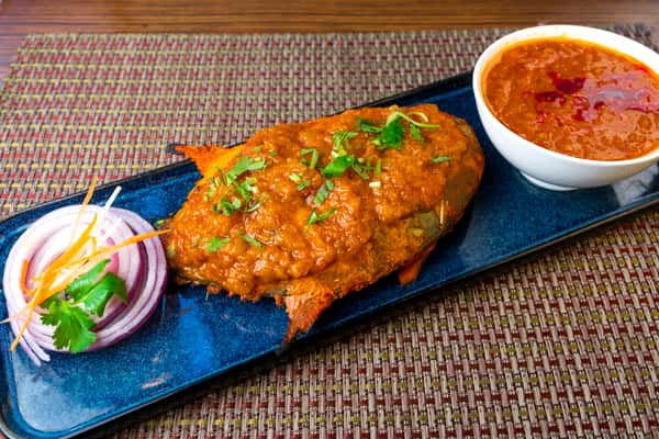 Whole fish topped with rich spiced masala gravy, served on a blue platter with onion slices and a side of curry sauce — traditional Indian seafood entrée.