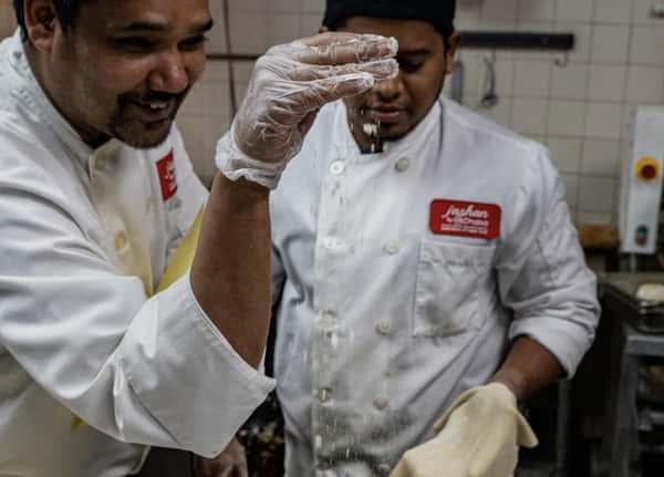 Chefs in a professional kitchen preparing fresh Indian cuisine, sprinkling ingredients by hand while shaping dough, highlighting authentic cooking techniques and teamwork.