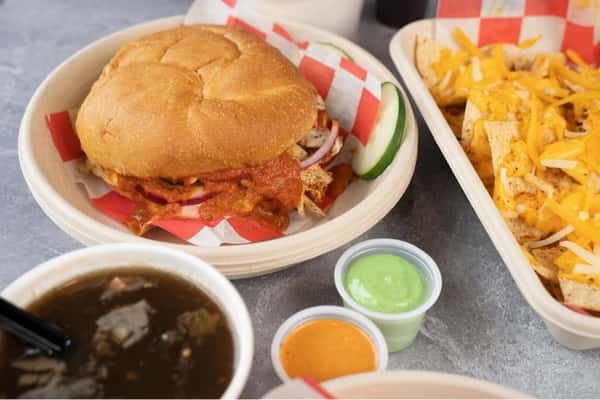Indian street food–inspired meal featuring a saucy chicken tikka sandwich on a soft bun, loaded masala nachos with melted cheese, tangy chutneys, and a side of spiced soup, served on a casual restaurant table.