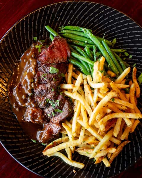 Medium-rare steak with peppercorn sauce, served with garlic herb fries and sautéed green beans on a black patterned plate.