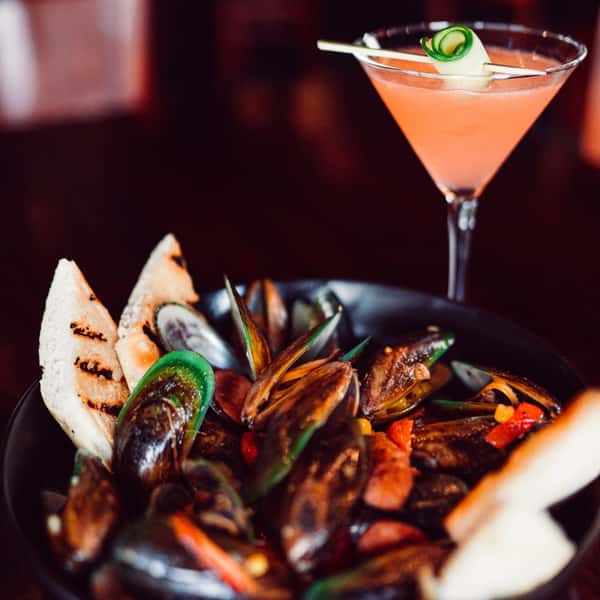 Bowl of steamed mussels with grilled bread and peppers, paired with a pink cocktail garnished with cucumber ribbon.