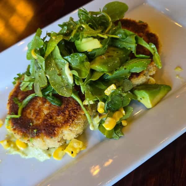 Golden crab cakes topped with arugula, avocado, corn, and cucumber salad on a creamy green herb sauce.