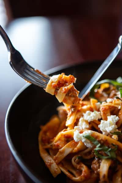 Close-up of tagliatelle pasta in rich tomato sauce with crumbled ricotta cheese and fresh basil, twirled on a fork.
